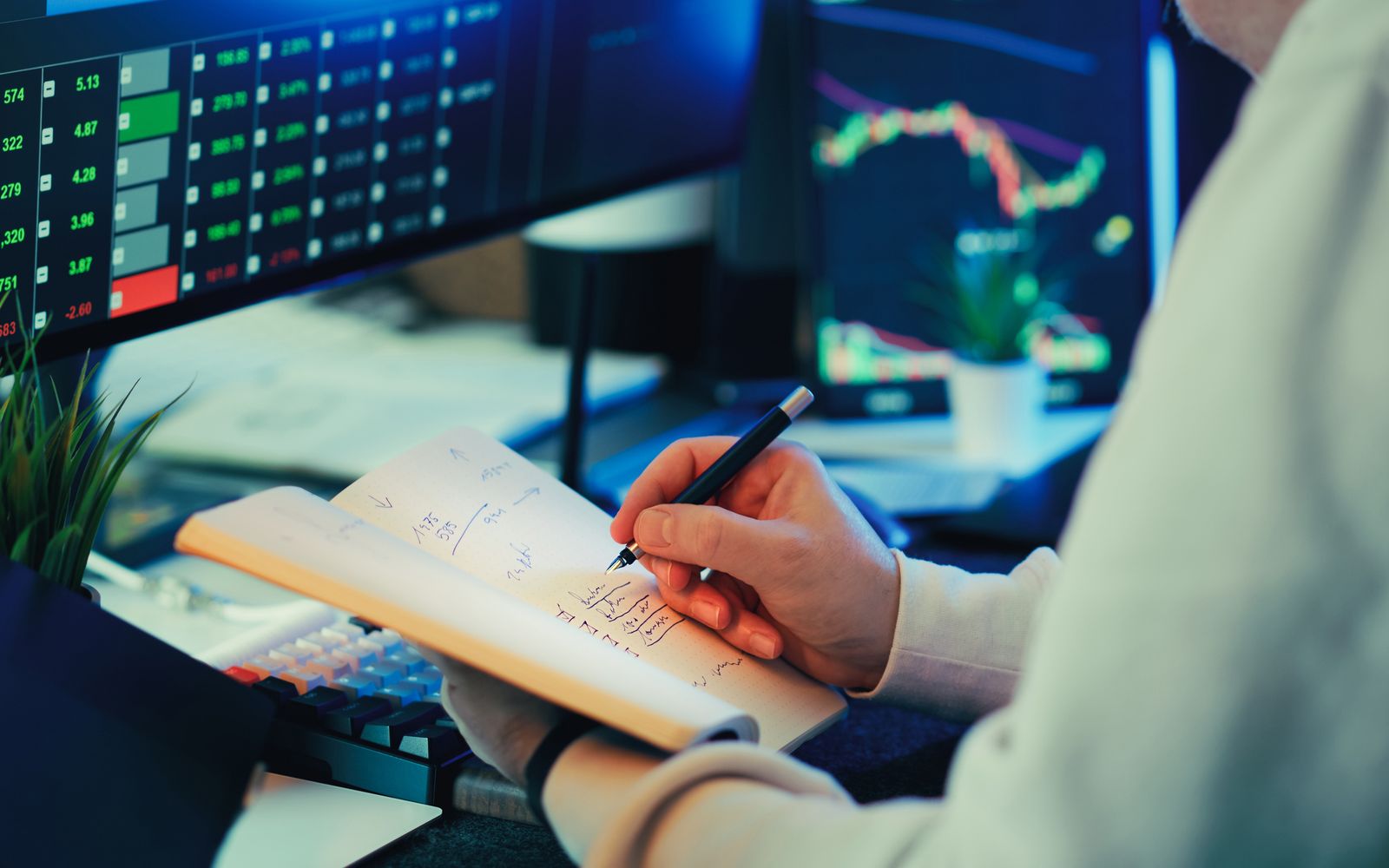Pessoa analisando dados financeiros em telas, fazendo anotações