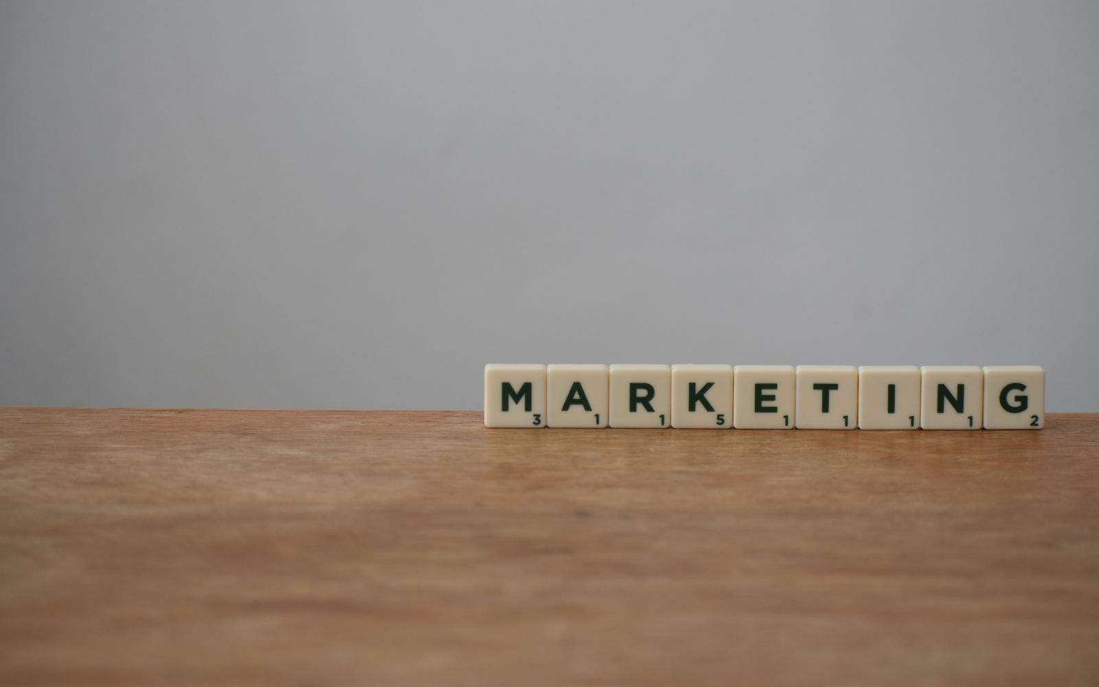 Peças do Scrabble formando a palavra MARKETING sobre uma mesa de madeira com fundo neutro