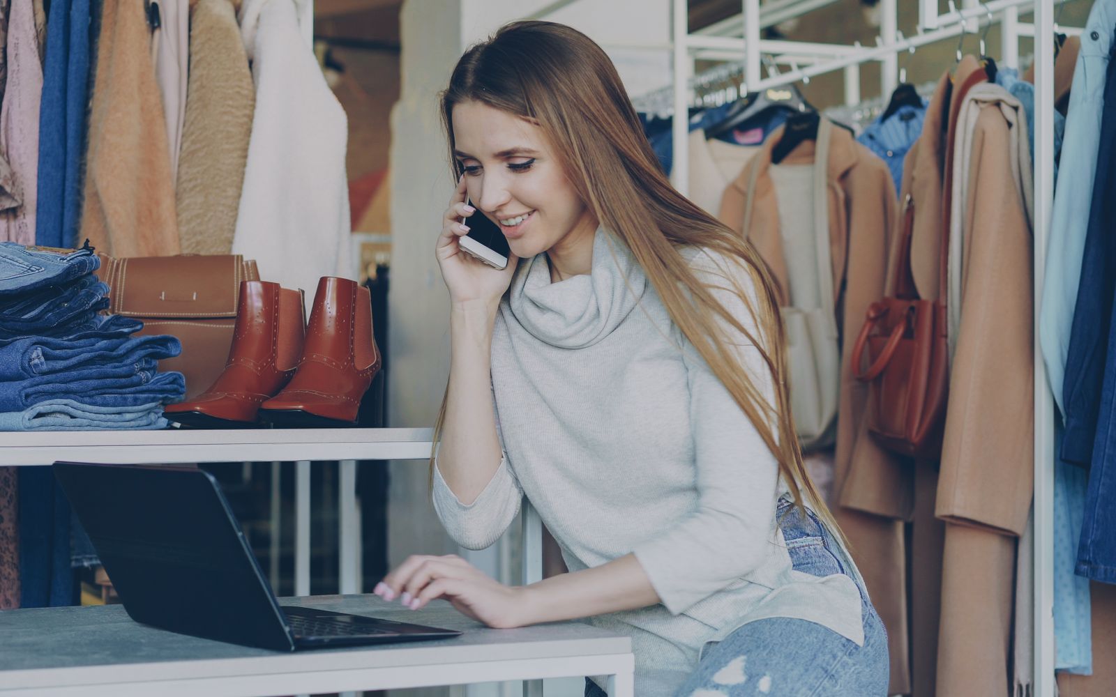 Mulher sorridente em uma boutique de moda usando um laptop e um celular