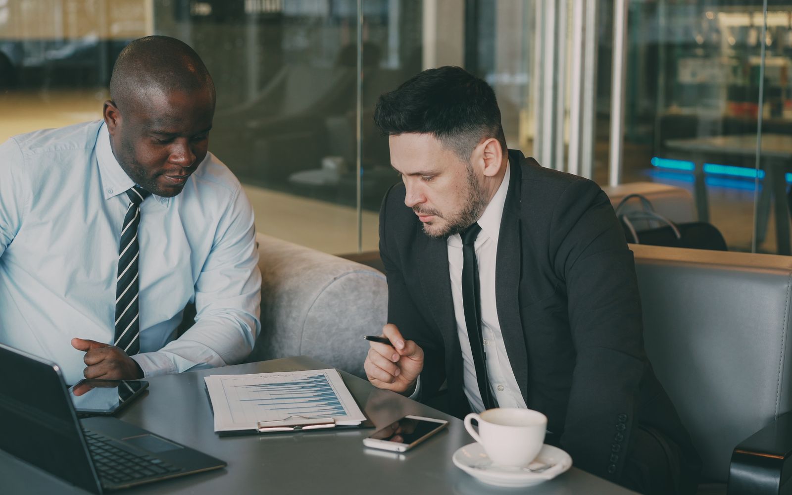 Dois homens de negócios revisando um relatório financeiro com laptop e café em ambiente de escritório