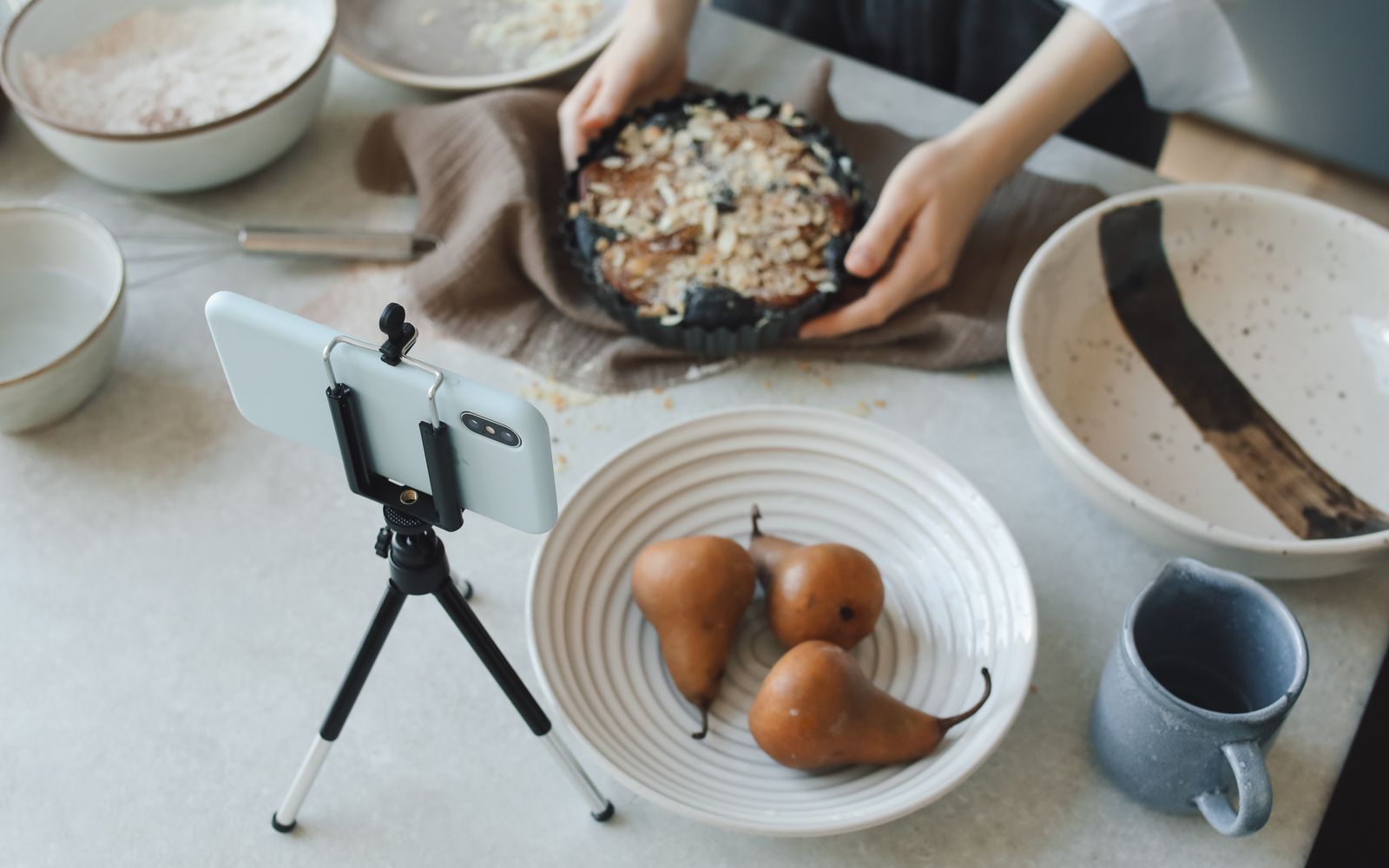 Mulher cozinhando enquanto grava com o celular, mostrando peras e um bolo