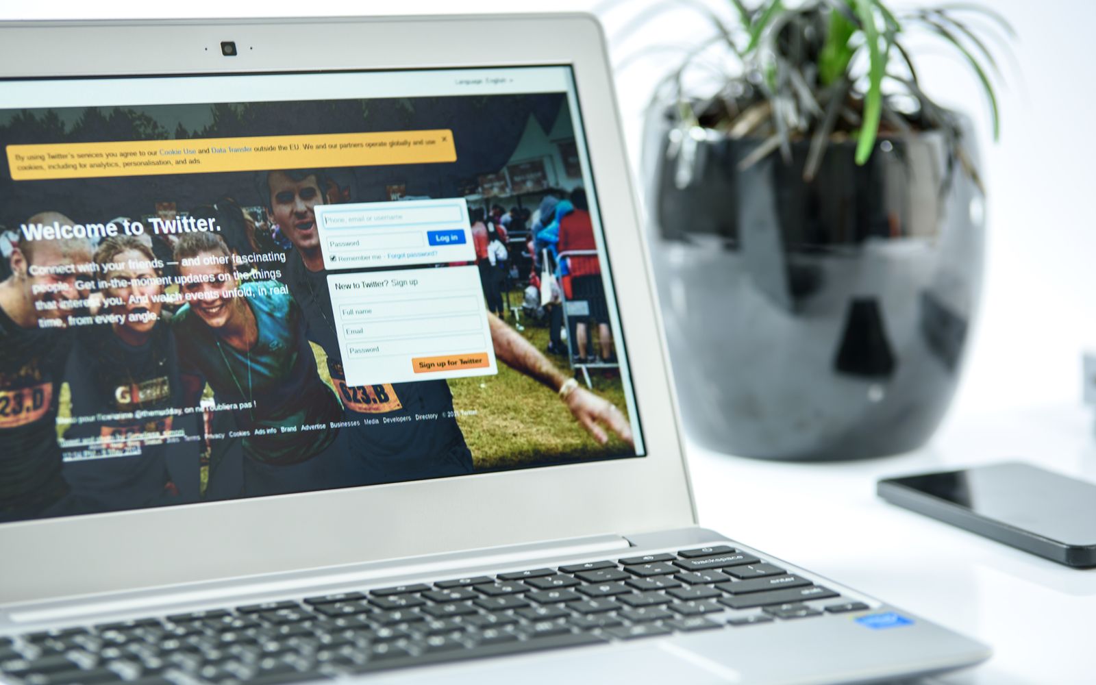Notebook exibindo a tela de login do Twitter, ao lado de um celular e um vaso