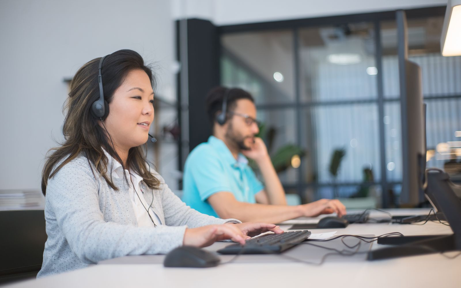 Dois profissionais de headset atendendo em computadores no escritório