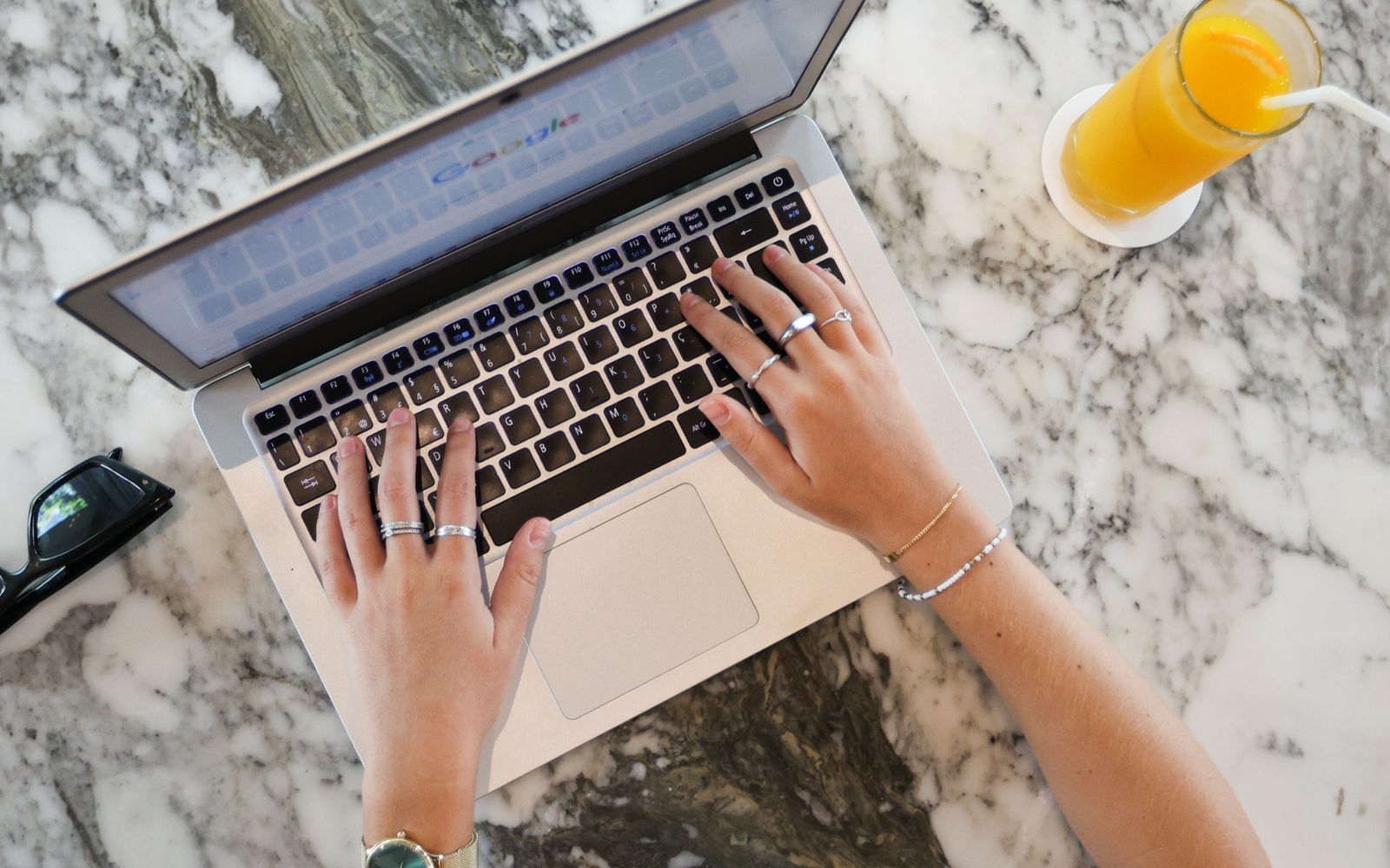 Mãos digitando em um notebook sobre bancada de mármore, com um copo de suco