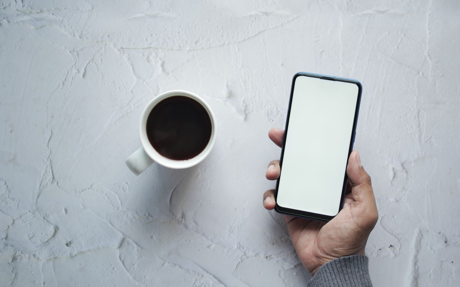 Mão segurando celular de tela apagada ao lado de uma xícara de café